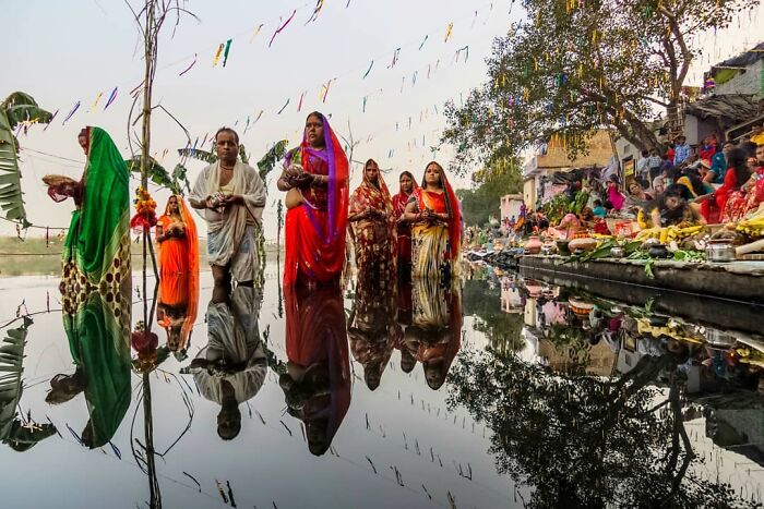 Women in colorful traditional attire standing and praying near a reflective water body, capturing everyday life in India.