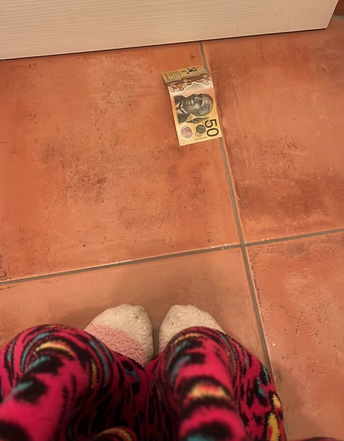 View of a child in colorful pajamas and fuzzy socks standing near a folded 50-dollar bill on a tiled floor, funny and wholesome moment.