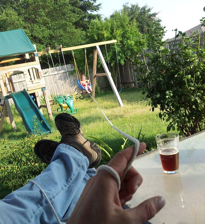 Dad relaxing with drink while casually pushing child on swing, showcasing funny differences between moms and dads in parenting.