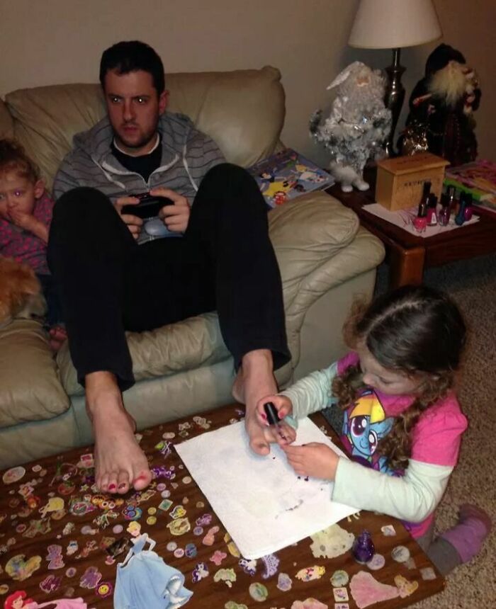 Man playing video games while daughter paints his toenails, showcasing funny photos revealing differences between moms and dads.