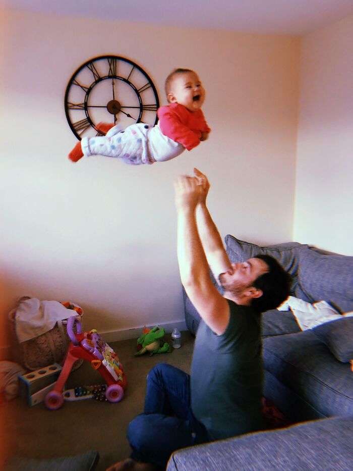 Dad joyfully tossing baby in the air in living room, showing funny differences between moms and dads in parenting moments.
