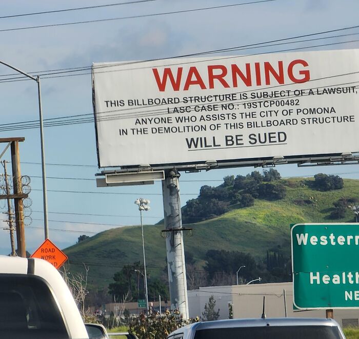 Funny and weird billboard warning about a lawsuit for demolition, standing tall against a green hillside and clear sky.