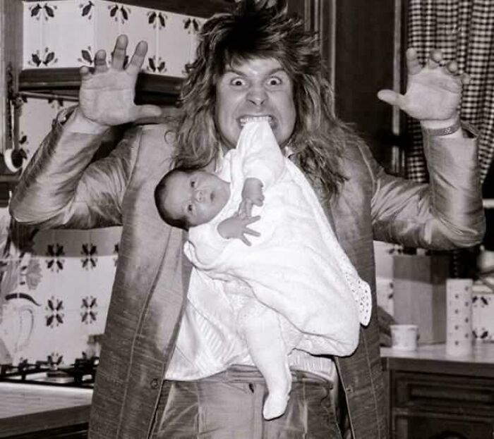 Man with wild hair holding a baby in a blanket in kitchen, showing playful expression, inspiring facts concept.