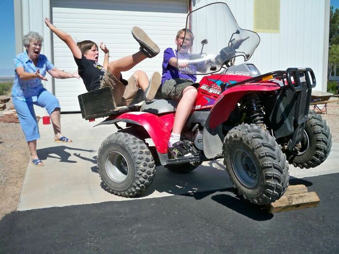 Two kids on an ATV with the front wheels lifted, a boy falling off and an older woman reaching out before disaster hits.
