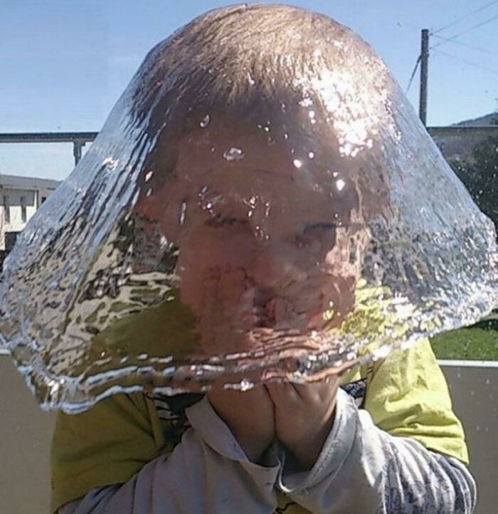Child caught in the split second before disaster as a large water balloon bursts over their head outdoors in bright sunlight