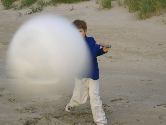 Young boy swinging baseball bat on sandy beach capturing split second before disaster hits with ball blurry and close.