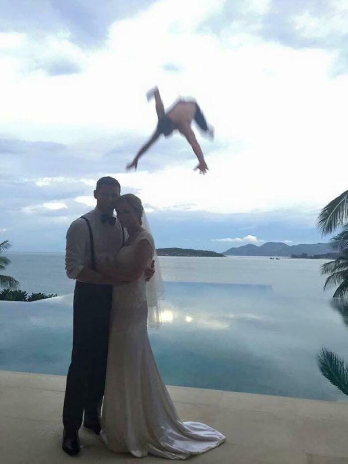 Bride and groom posing by infinity pool with person mid-air about to land in a hilarious split second before disaster moment