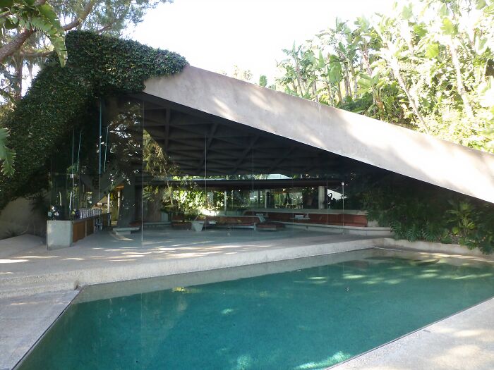 Modern iconic TV house with glass walls and a slanted roof next to a swimming pool in a lush outdoor setting.