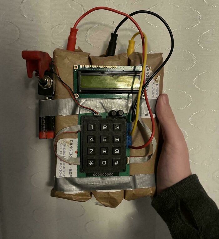 Hand holding a homemade electronic device with wiring, keypad, and taped components showing poor electronics handling.
