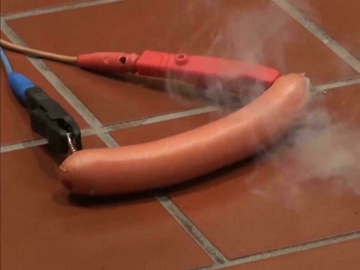 Hot dog connected to electrical clamps with smoke rising, illustrating mishandling of electronics and soldering hazards.