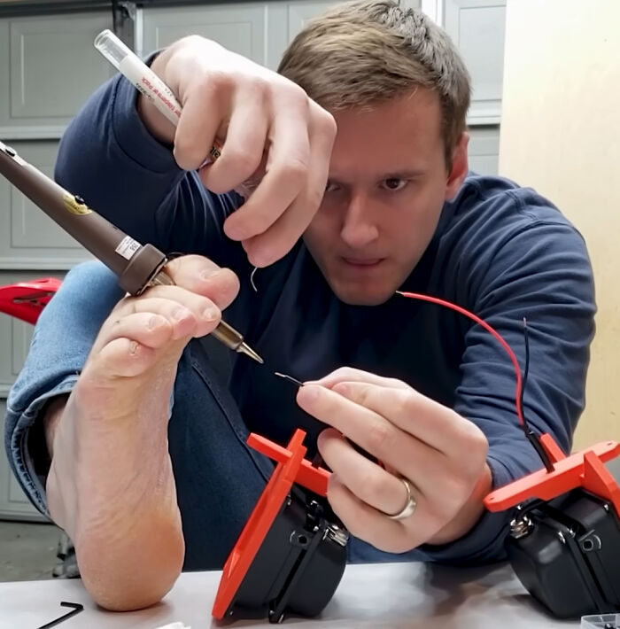 Man using foot to hold soldering iron while attempting to solder wires in an unconventional electronics handling method.