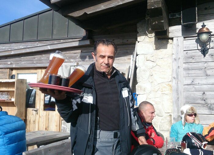 Man carrying tray with several glasses of beer outside a wooden cabin, moments before a likely spill disaster.