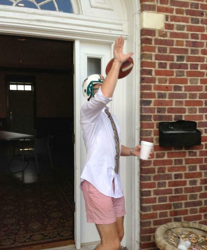 Man in helmet and tie attempting to catch football with cup in hand, capturing split second before disaster.