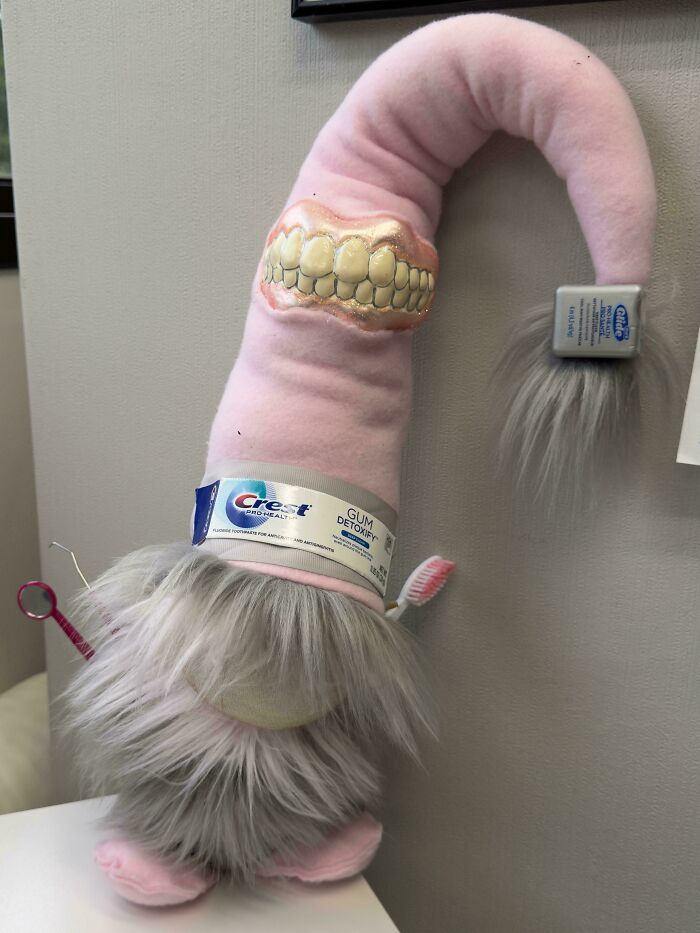 Weird plush character with realistic teeth, pink body, gray fur, and Crest toothpaste tube wrapped around it.
