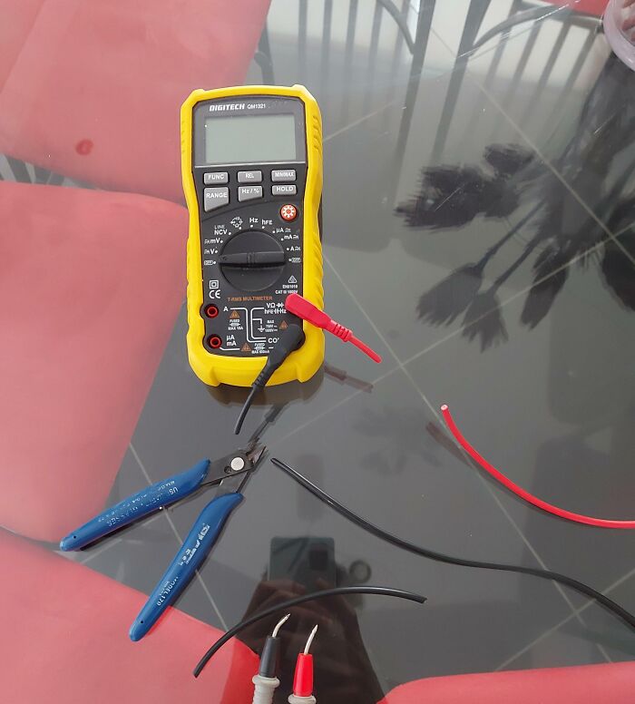 Digital multimeter and wire cutters on glass table with stripped wires showing common electronics handling tools.