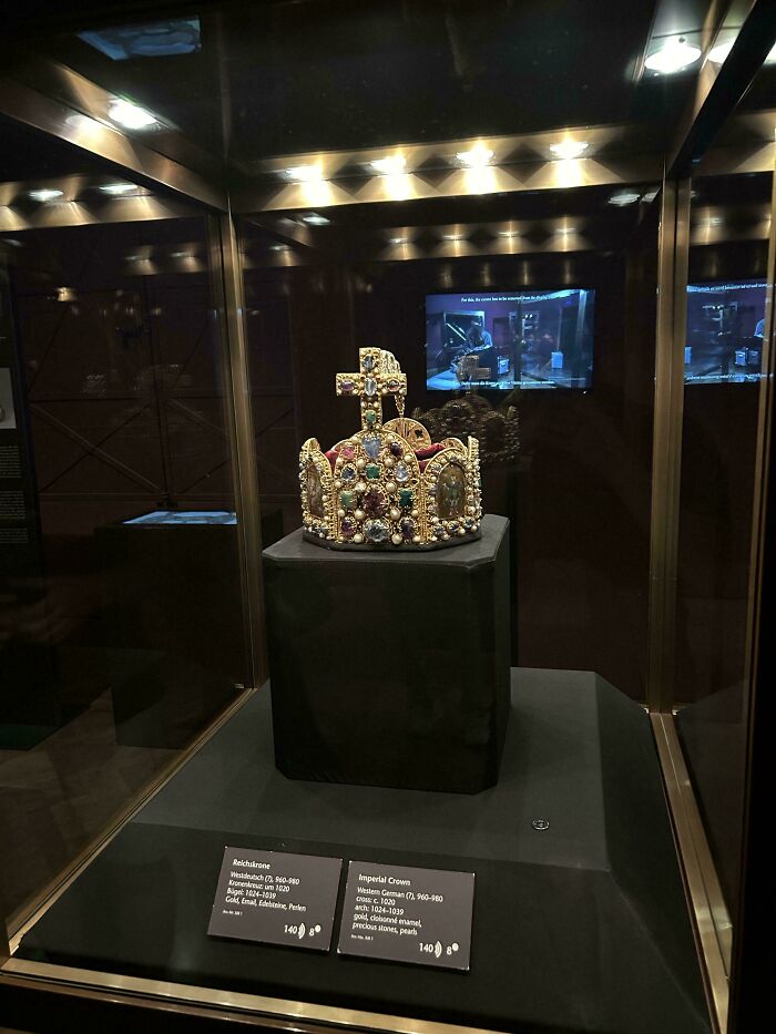 Medieval history enthusiasts exploring an illuminated medieval crown with gold, precious stones, and pearls in a museum display.