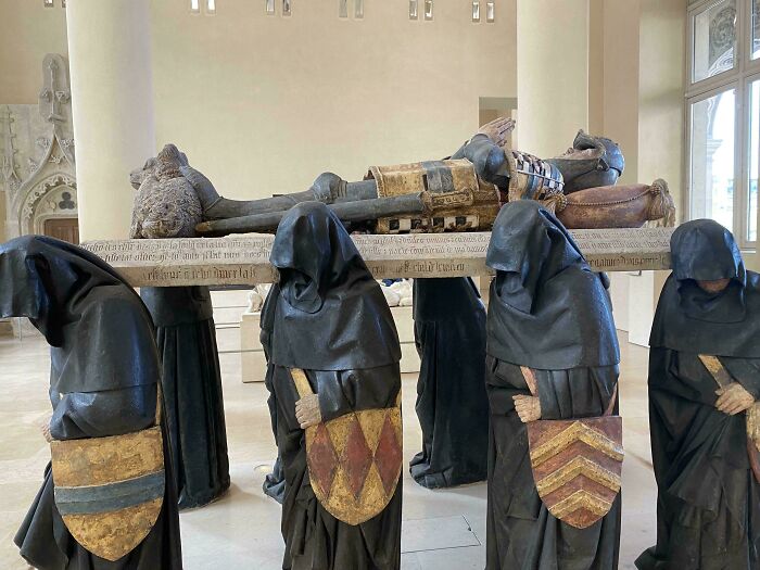 Medieval history enthusiasts appreciate intricate hooded statues carrying a knight effigy in a historic museum setting.