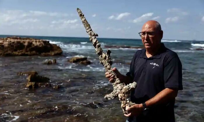 Man holding a barnacle-covered medieval sword by the rocky shore, reflecting medieval history enthusiasts' recent joy.