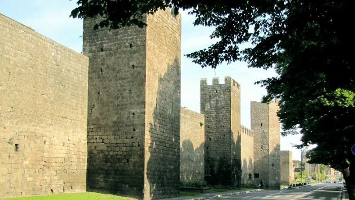Medieval history enthusiasts explore tall stone fortress walls and towers on a sunny day with tree shadows nearby.
