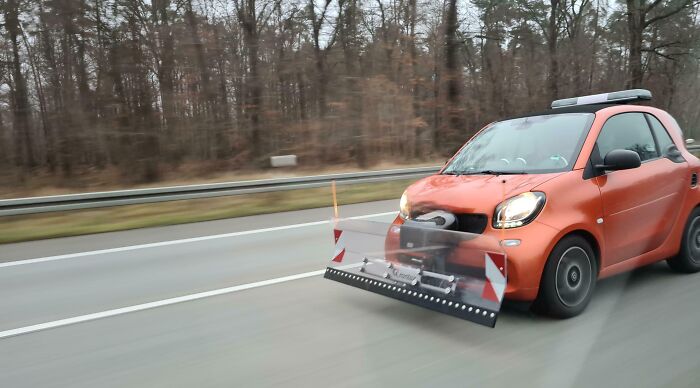 Small orange car with an unusual front attachment driving on a highway in a forested area, weird and cursed image.
