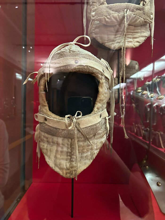 Medieval history quilted arming cap displayed in a museum case with a red background and armor pieces nearby.