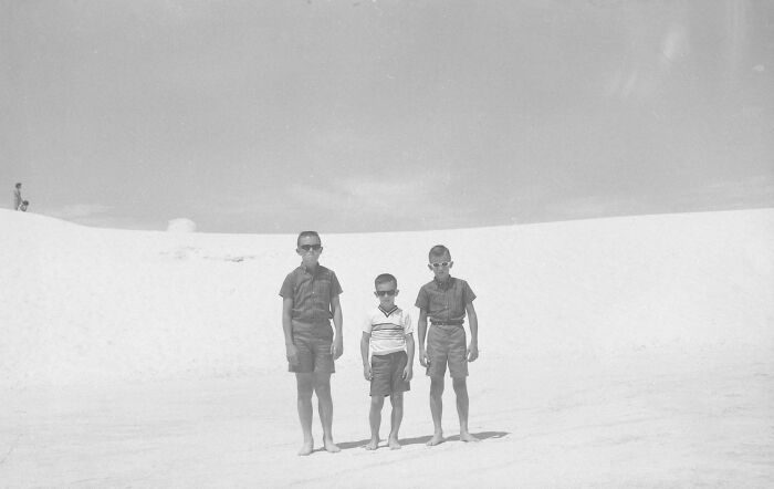 Three boys wearing sunglasses standing barefoot on sand in a vintage vacation pic from a time before cell phone cameras.