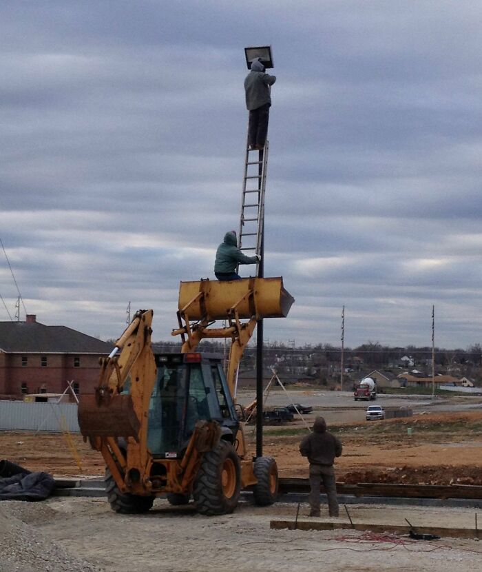 Construction workers using a backhoe to hold a ladder with a worker fixing a light, showing unsafe job conditions.