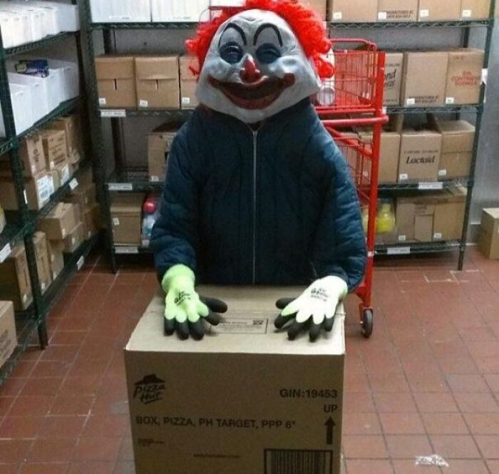 Warehouse worker wearing a creepy clown mask and gloves standing behind a cardboard box in storage, unsettling job photo.