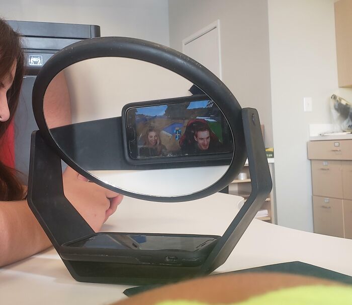 Kid using a homemade invention to display a phone elevated on a unique mirror stand on a table indoors.