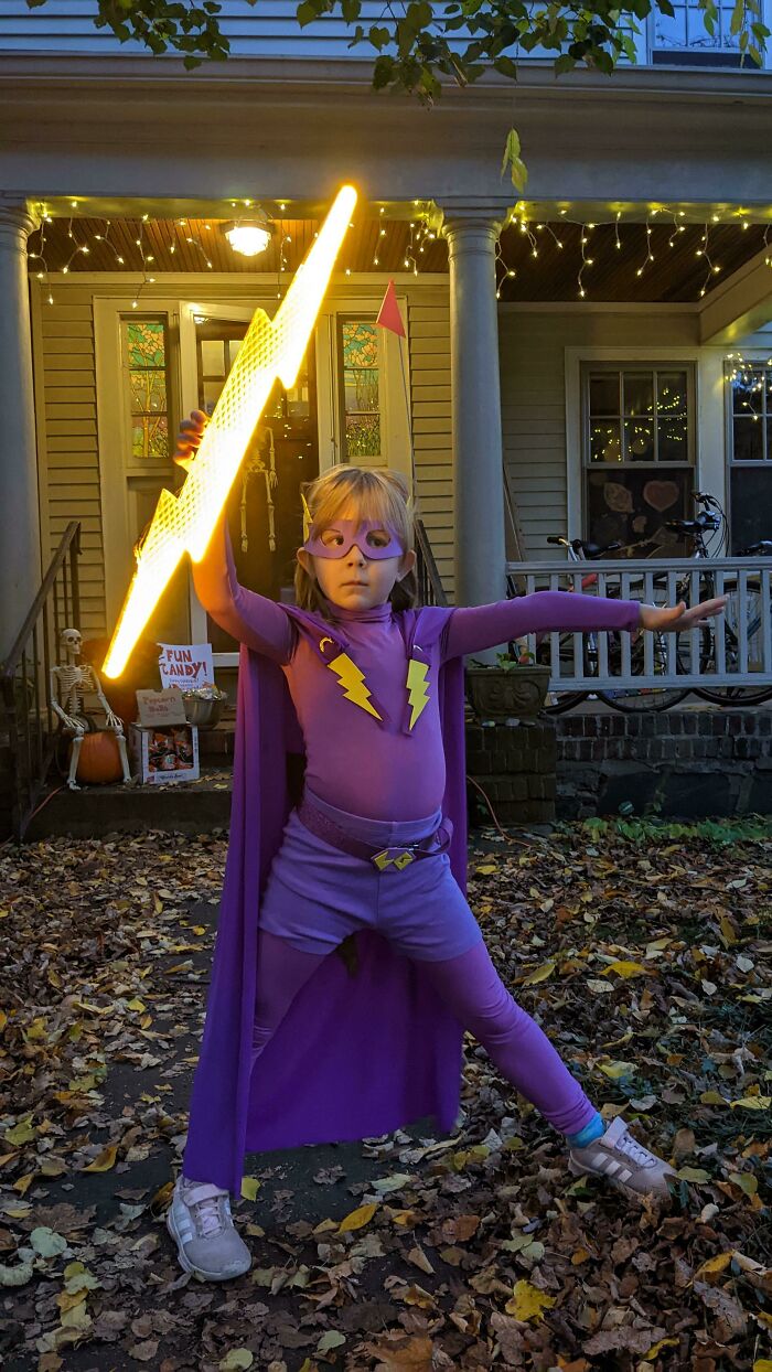 Child dressed as superhero holding glowing lightning bolt invention outside house at night among fallen leaves.