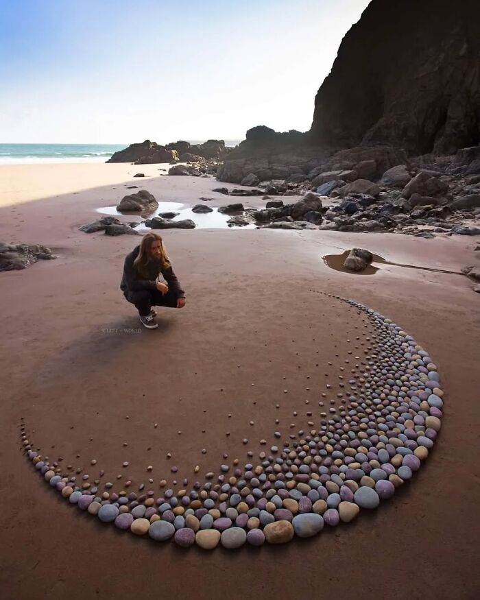 Man crouching by a crescent shaped stone sculpture on a sandy beach, part of Jon Foreman's hypnotic land art in Wales.