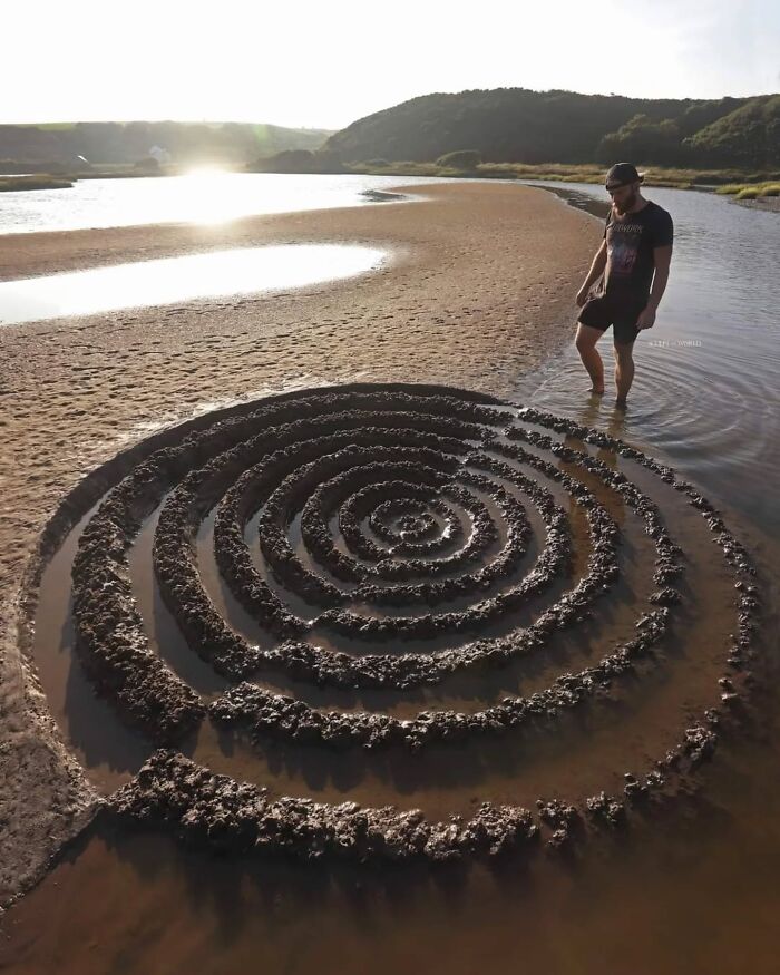 Man standing in shallow water next to hypnotic spiral land art sculpted on a sandy beach in Wales.