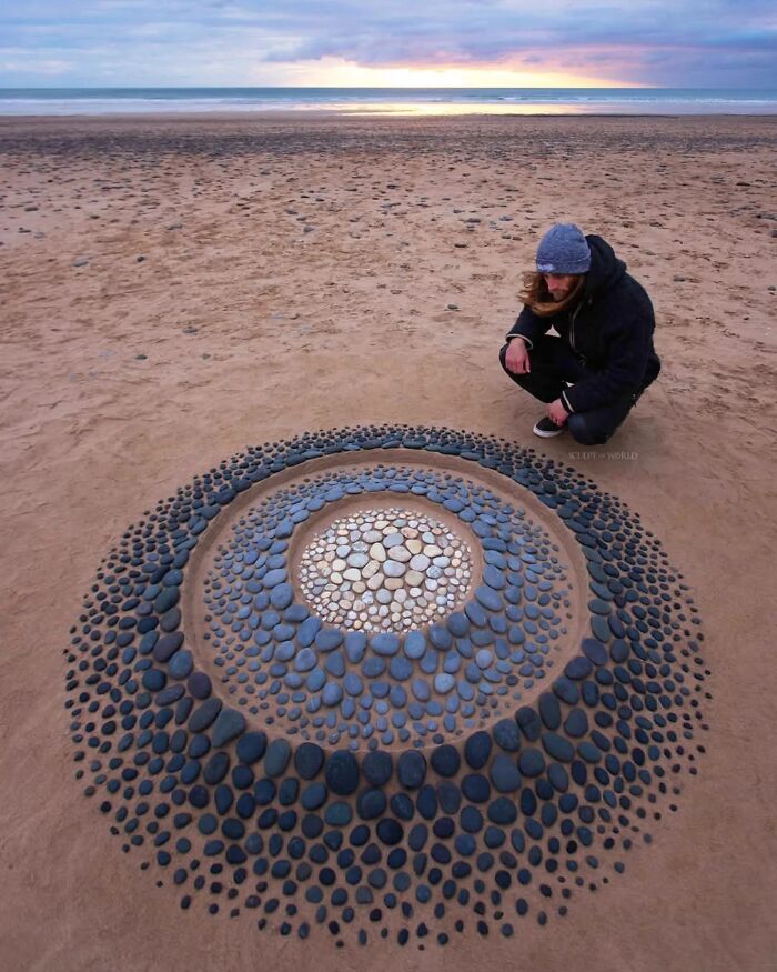 Man crouching beside hypnotic land art made of concentric circles of stones on a sandy Wales beach at sunset.