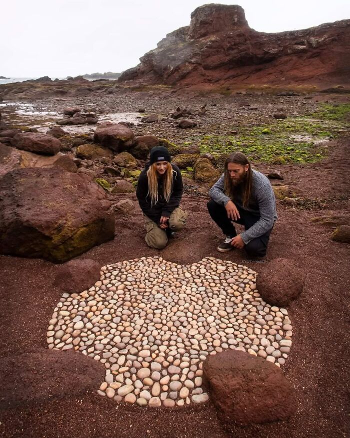 Two artists sculpt land art with smooth stones on Wales’s rocky beach surrounded by large red rocks and green algae.