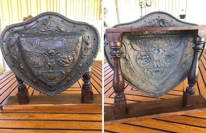 Ornate old shield-shaped object with detailed floral and animal engravings on a wooden table outdoors.