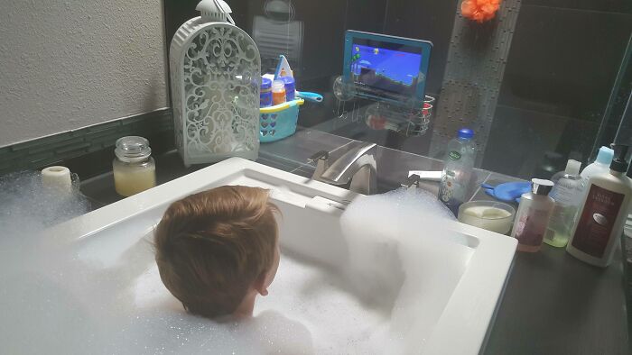 Child in bubble bath watching tablet mounted near faucet, showcasing kids inventions surprising parents with smart setup.