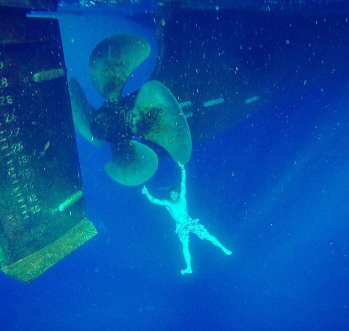 Diver underwater holding onto a massive ship propeller, demonstrating human for scale in deep blue ocean.