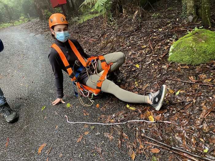 Person wearing orange safety gear and helmet sitting on forest path next to a large earthworm for scale comparison.
