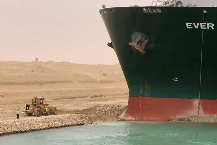 Massive cargo ship next to workers and a bulldozer showing human for scale in a sandy canal environment.