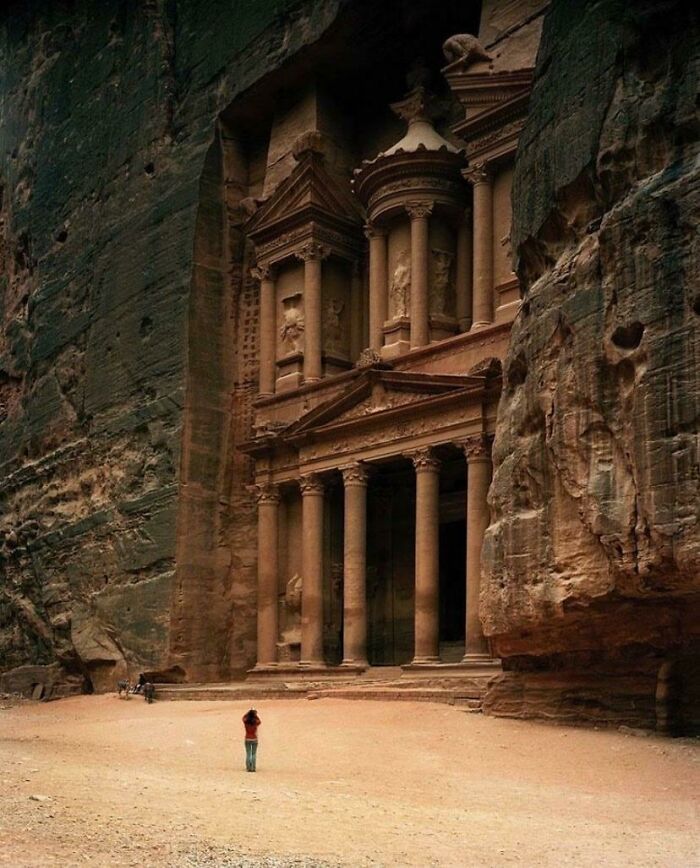 Person standing in front of the massive ancient rock-cut facade showing human for scale in a desert setting.