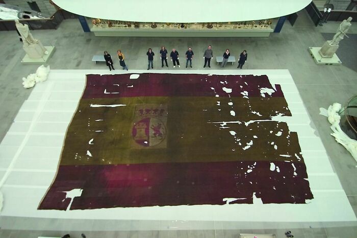 Group of people standing behind an enormous tattered flag displayed on the floor for human for scale comparison.