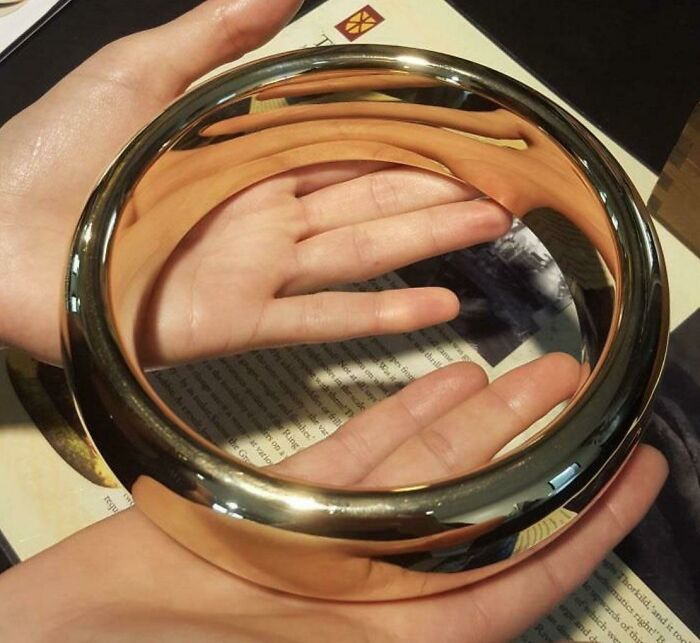 Hands holding a reflective metallic torus displaying human for scale with distorted reflections on its surface