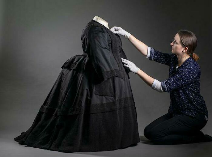 A woman wearing white gloves kneels adjusting a large black Victorian dress, showing human for scale and size comparison.