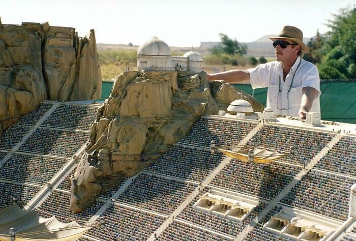 Man for scale standing next to a large detailed model of a stadium and rocky structure outdoors in sunlight