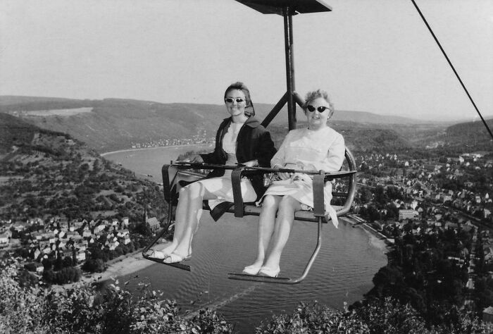 Two women wearing sunglasses on a ski lift overlooking a river and village in a vintage vacation photo.