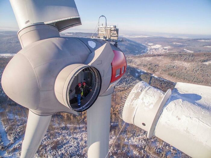 Two people inside the huge wind turbine hub showing human for scale against snowy forest landscape below.