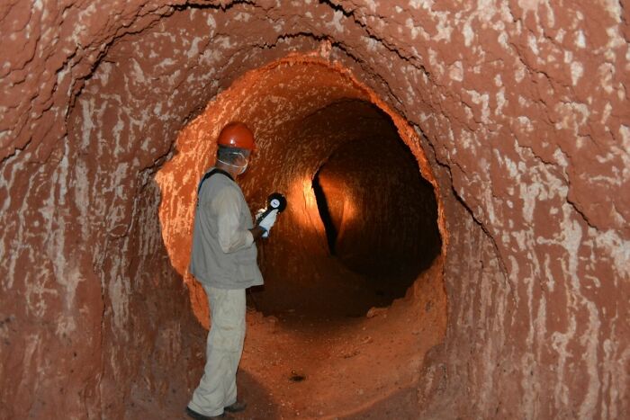 Person in protective gear holding a gauge inside a large tunnel showing human for scale in an unusual way.