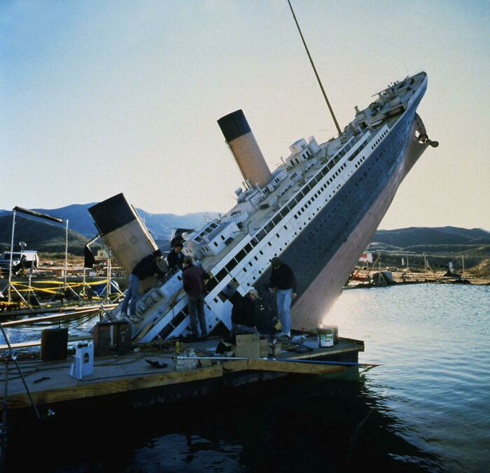 Workers on a dock next to a tilted ship model, showing human for scale and the unusual size comparison.