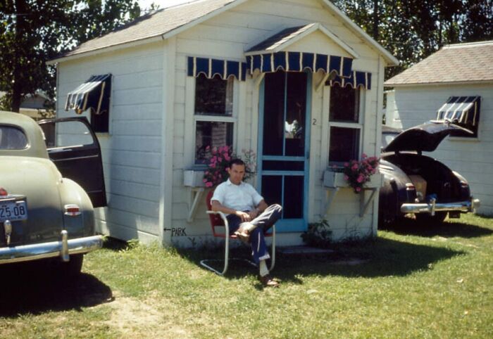 Man sitting in chair outside small vacation cabin with vintage cars, capturing vacation pics before cell phones had cameras.