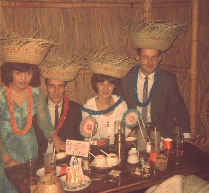 Vintage vacation photo of four people wearing Hawaii Kai hats and leis, enjoying a festive meal at a tiki-style table.